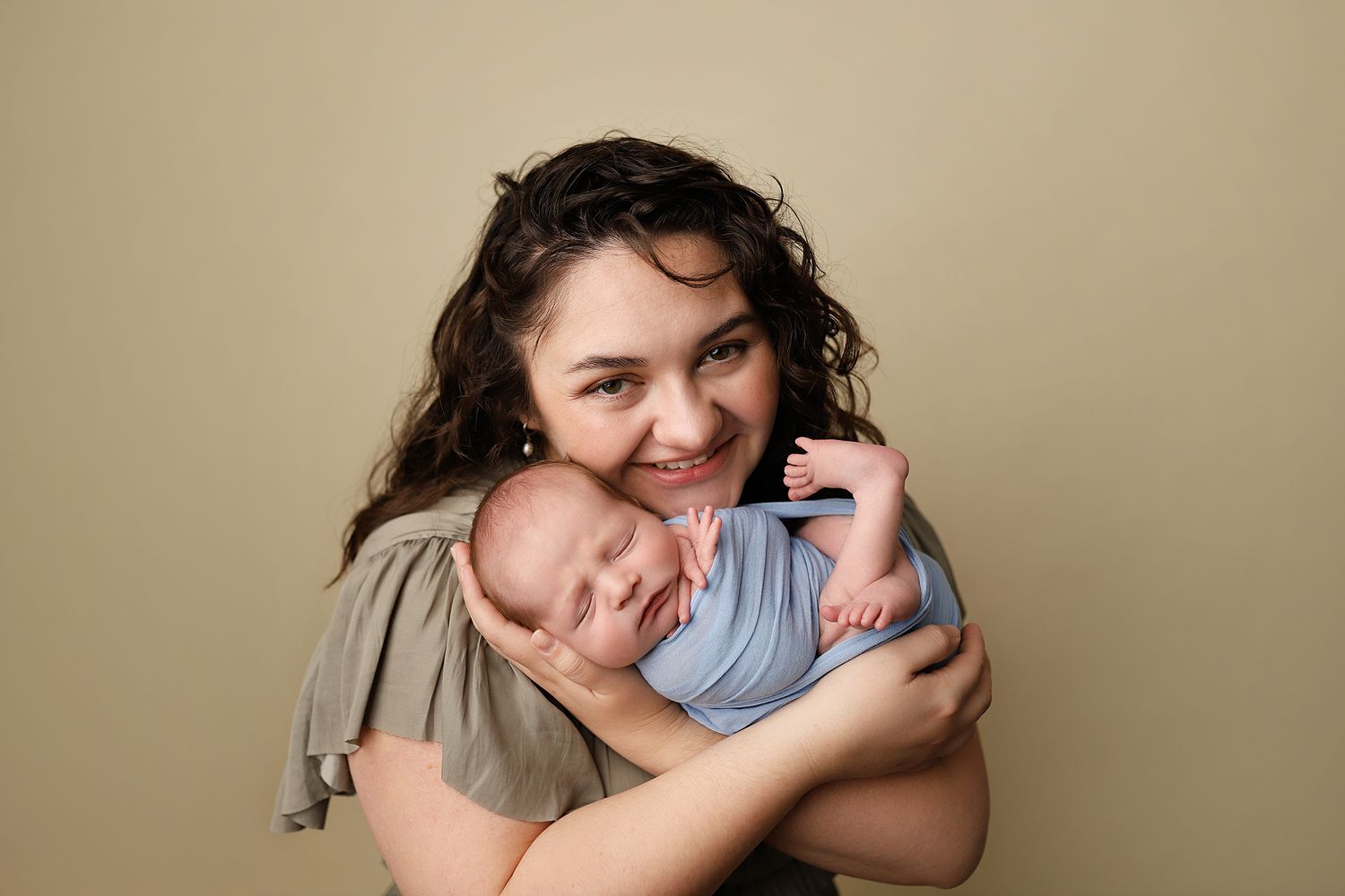 Smiling mother holding sleeping newborn baby wrapped in blue swaddle against neutral green background. Orem newborn photographer