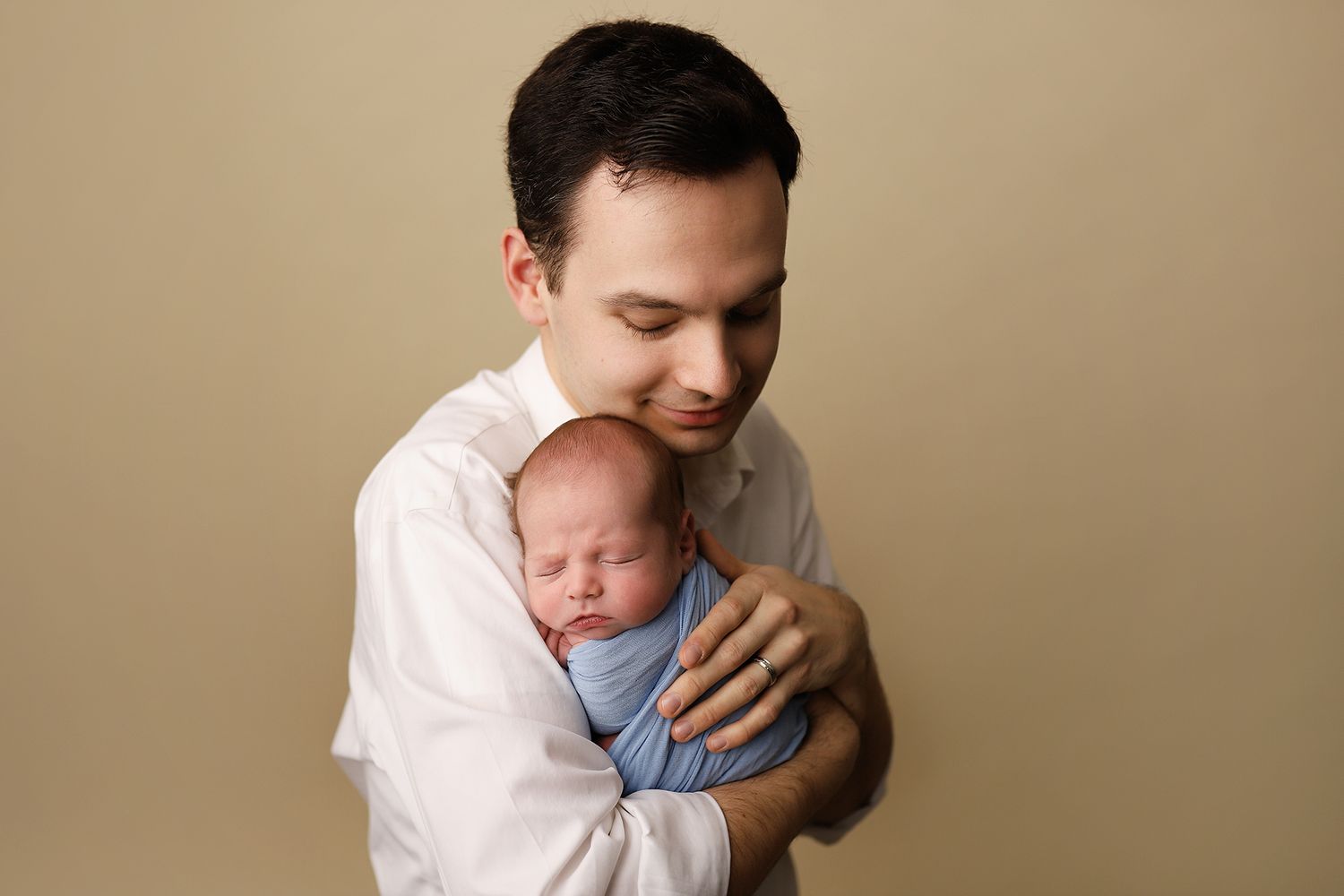 Father in white shirt tenderly holds sleeping newborn baby wrapped in blue against beige studio background. baby photographer in Utah County
