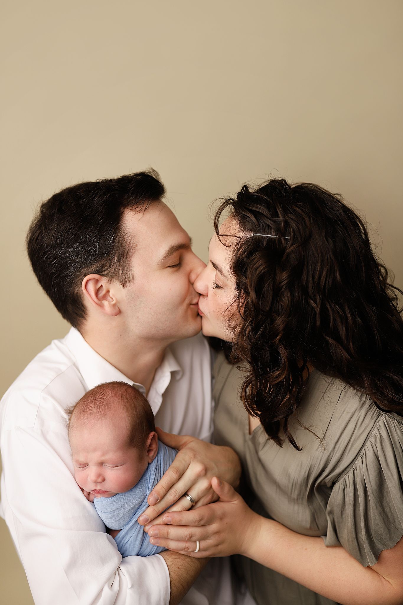 A couple shares a tender kiss while cradling their sleeping newborn baby wrapped in a blue swaddle.