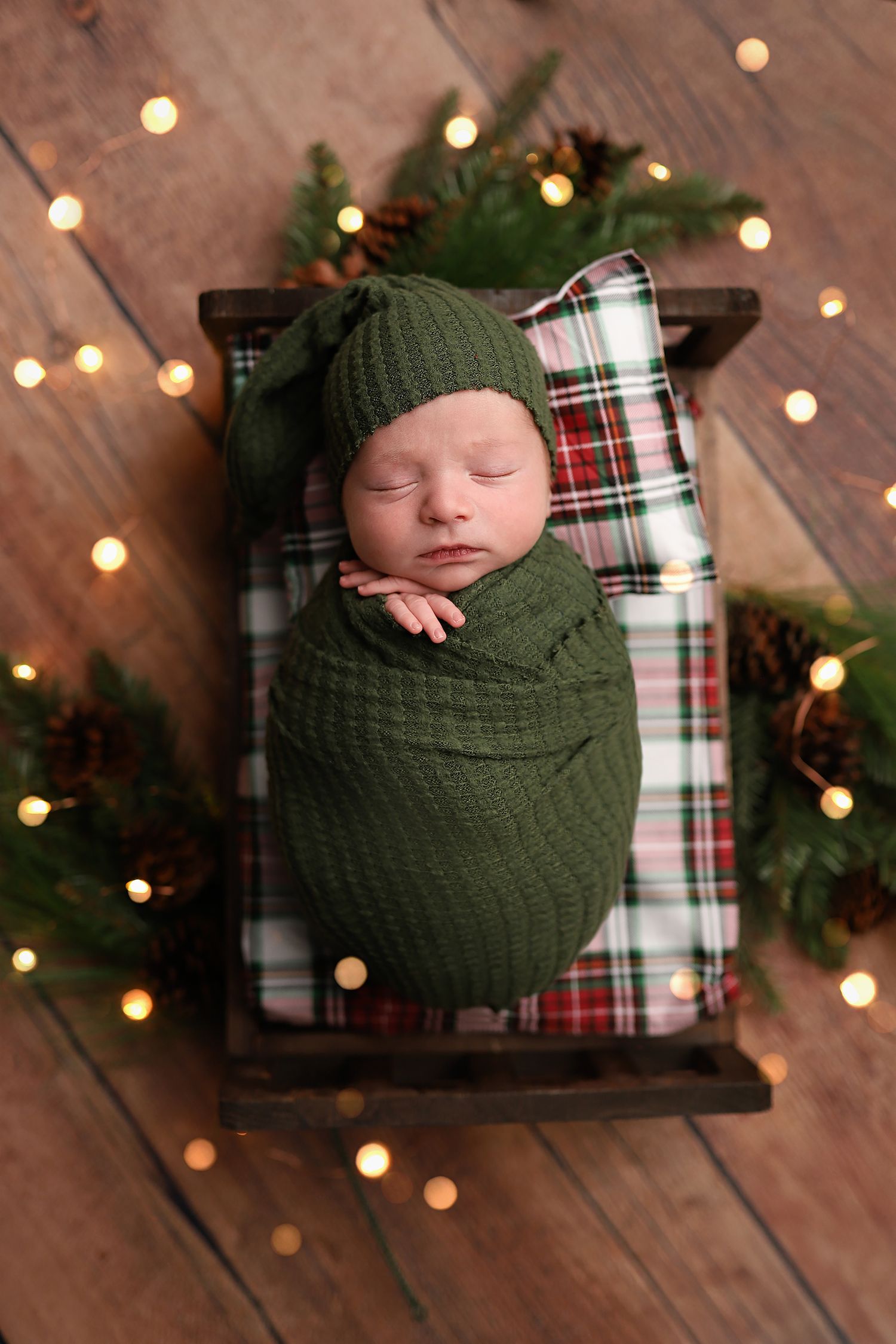 Newborn baby swaddled in green wrap and hat, sleeping in a mini wooden bed with plaid pillow and Christmas lights.