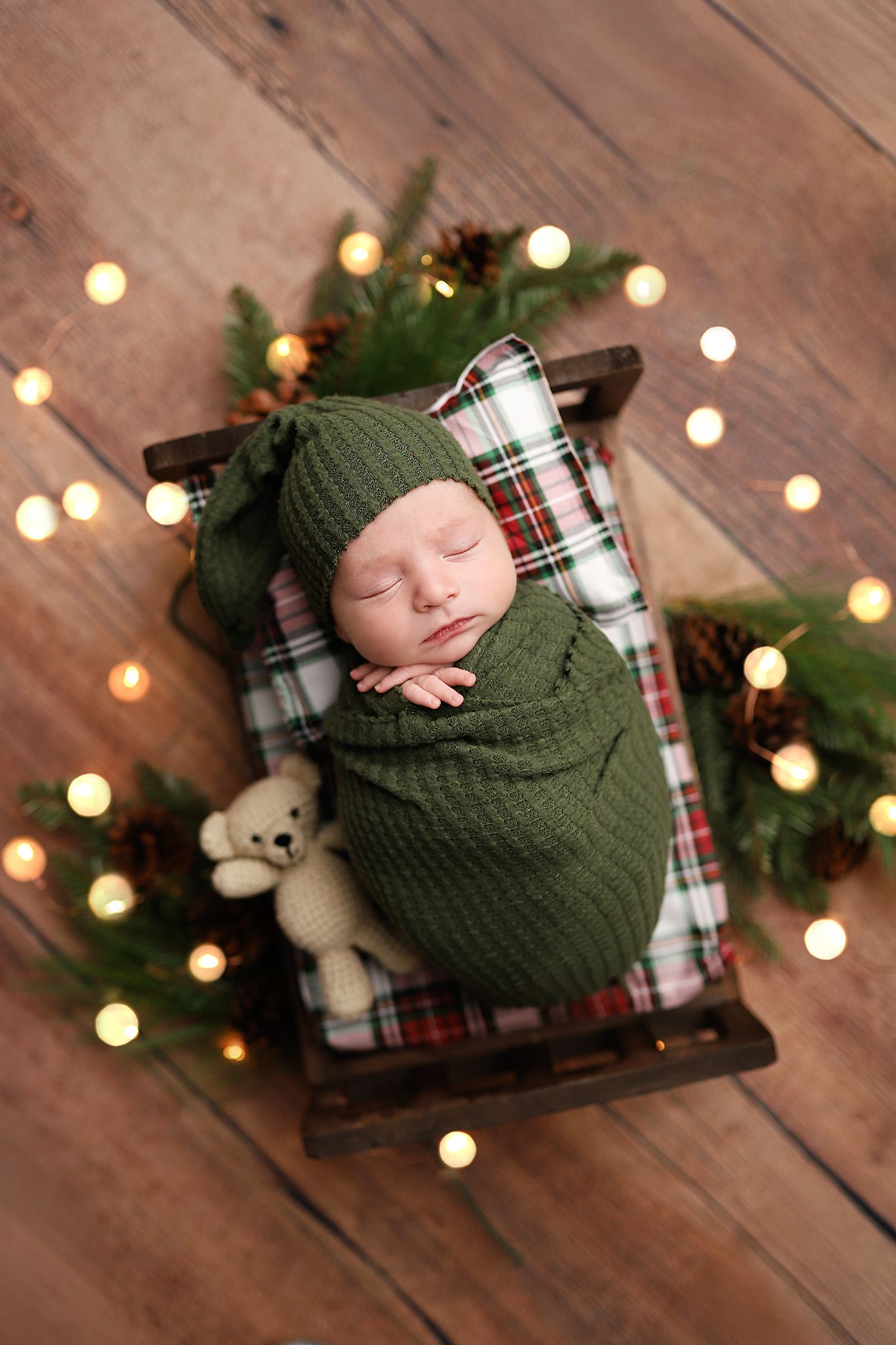 Newborn baby swaddled in green knit wrap with matching hat, sleeping in a miniature wooden bed with Christmas decorations.