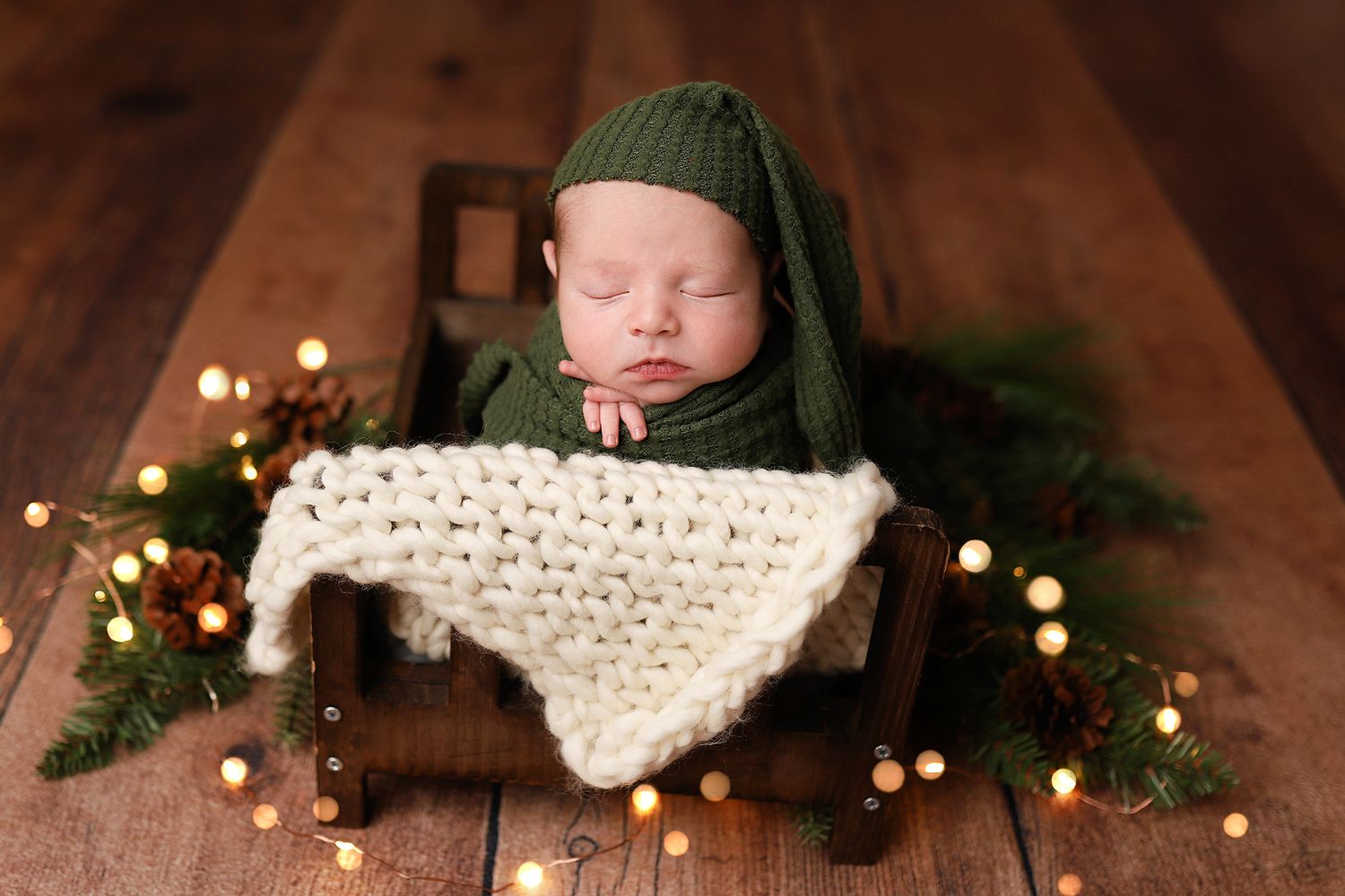Sleeping newborn in green knit hat nestled in a wooden crib with cream blanket, surrounded by festive holiday lights.