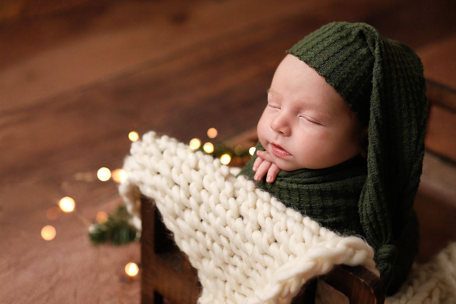 Peaceful newborn baby wearing a green knit hat, sleeping in a wooden crib with a cream blanket and bokeh lights.