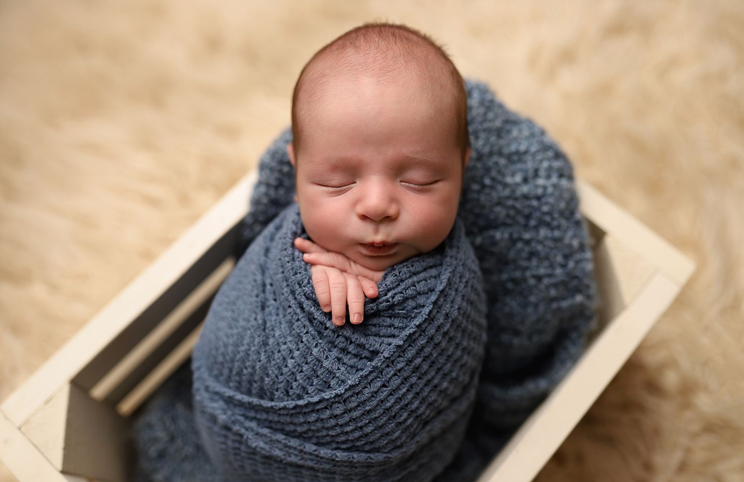 Newborn baby swaddled in a blue knit wrap, sleeping peacefully inside a small wooden crate on a soft beige background.