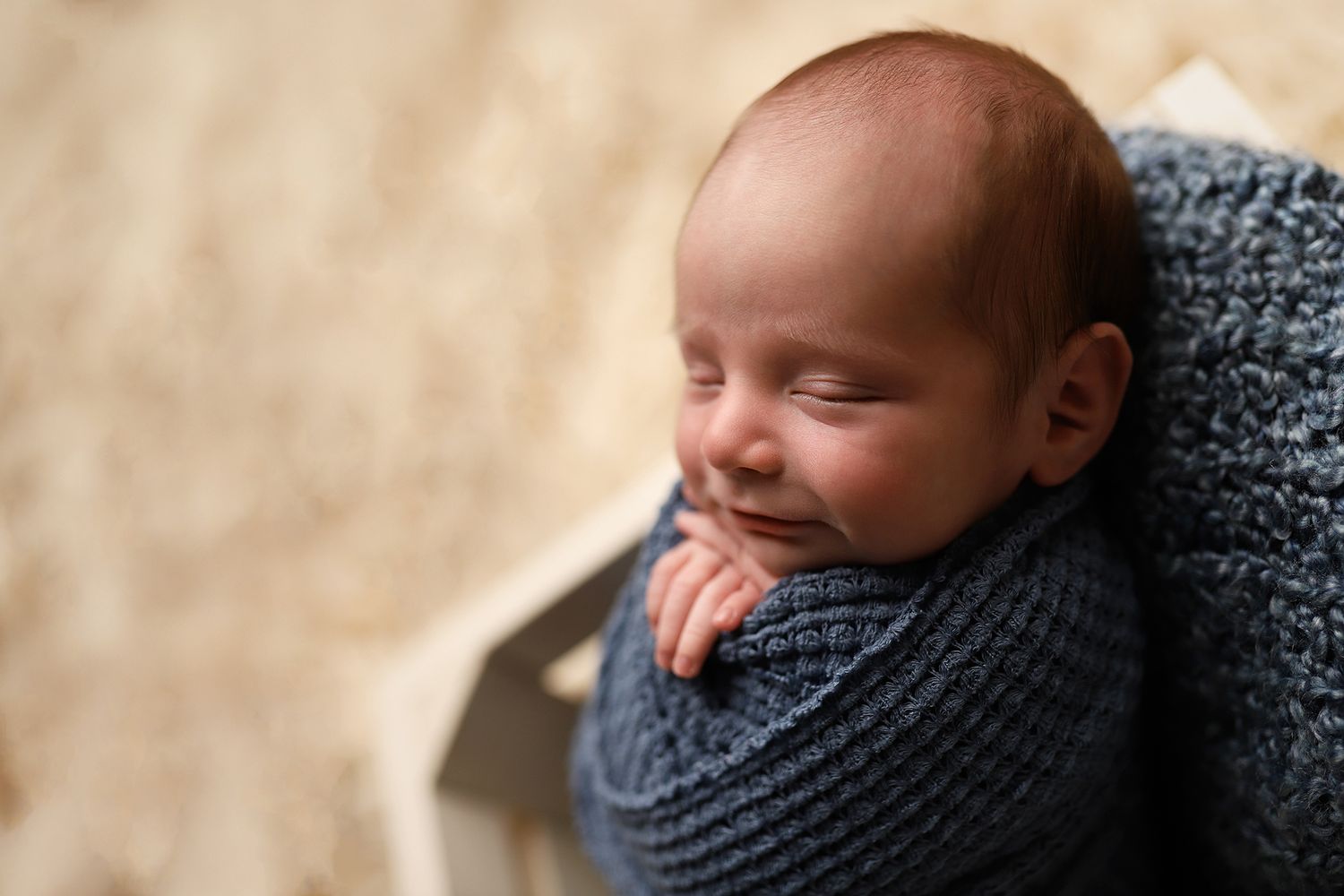 Peaceful newborn baby swaddled in a soft blue knit wrap, sleeping with a gentle smile in a wooden box. Utah newborn photographer