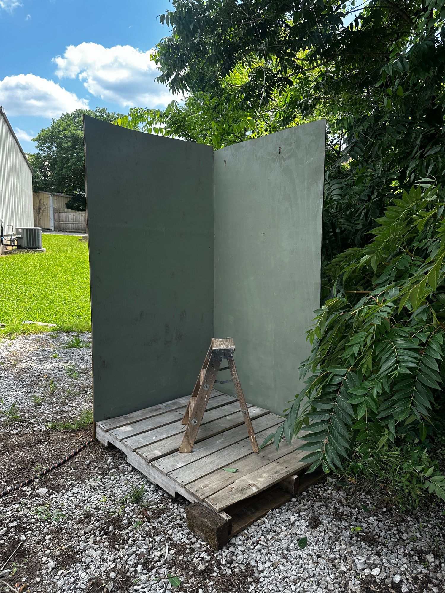 Outdoor green painted wall panels on wooden pallets with a small ladder, surrounded by gravel and lush trees.