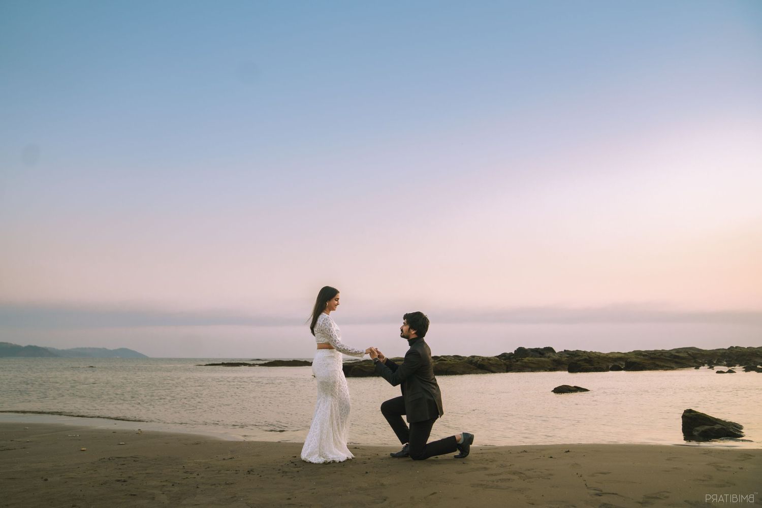 Man proposing on one knee to woman in white dress on sandy beach at sunset with ocean backdrop.