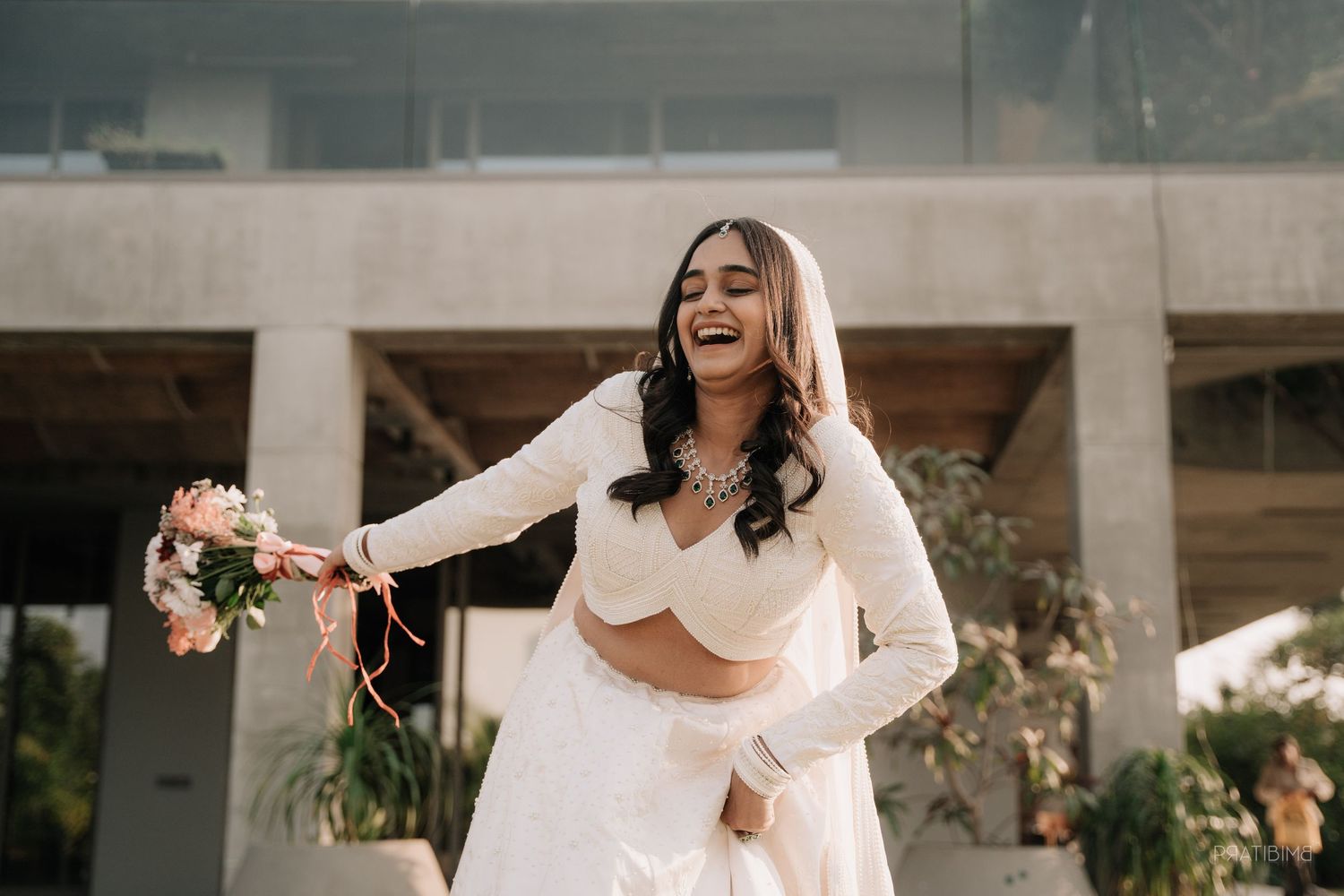 Joyful bride in white lehenga laughing and holding a pink bouquet outdoors at a modern venue.
