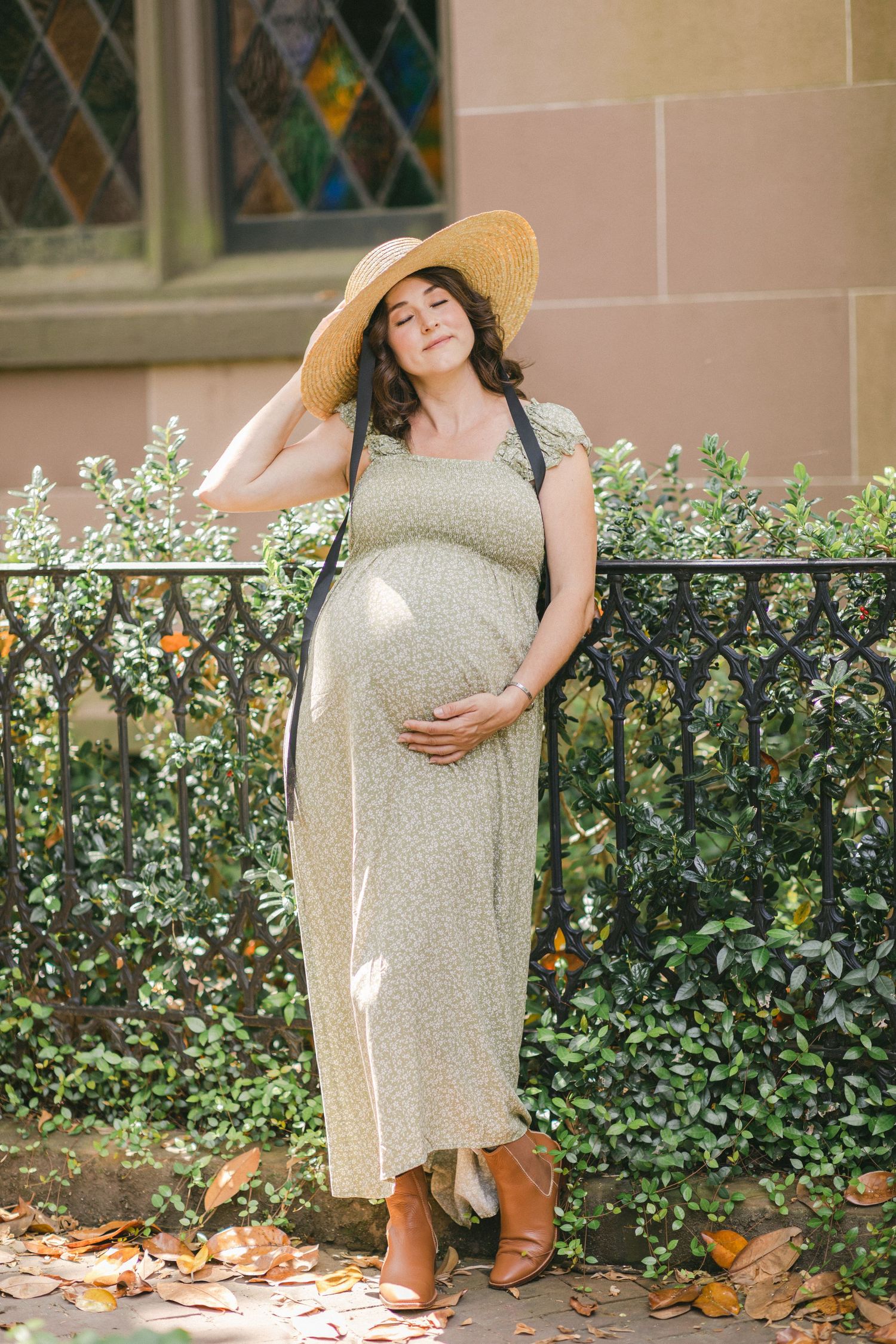 Pregnant woman in green midi dress and straw hat poses outdoors by iron fence with lush greenery.