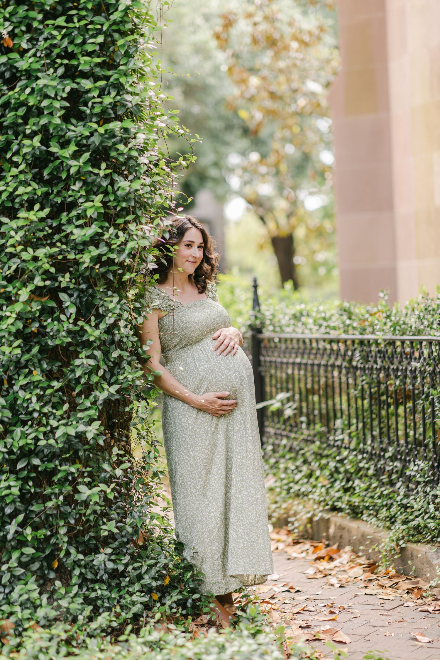 Pregnant woman in sage green dress cradling her belly outdoors near lush green hedges and iron fence.
