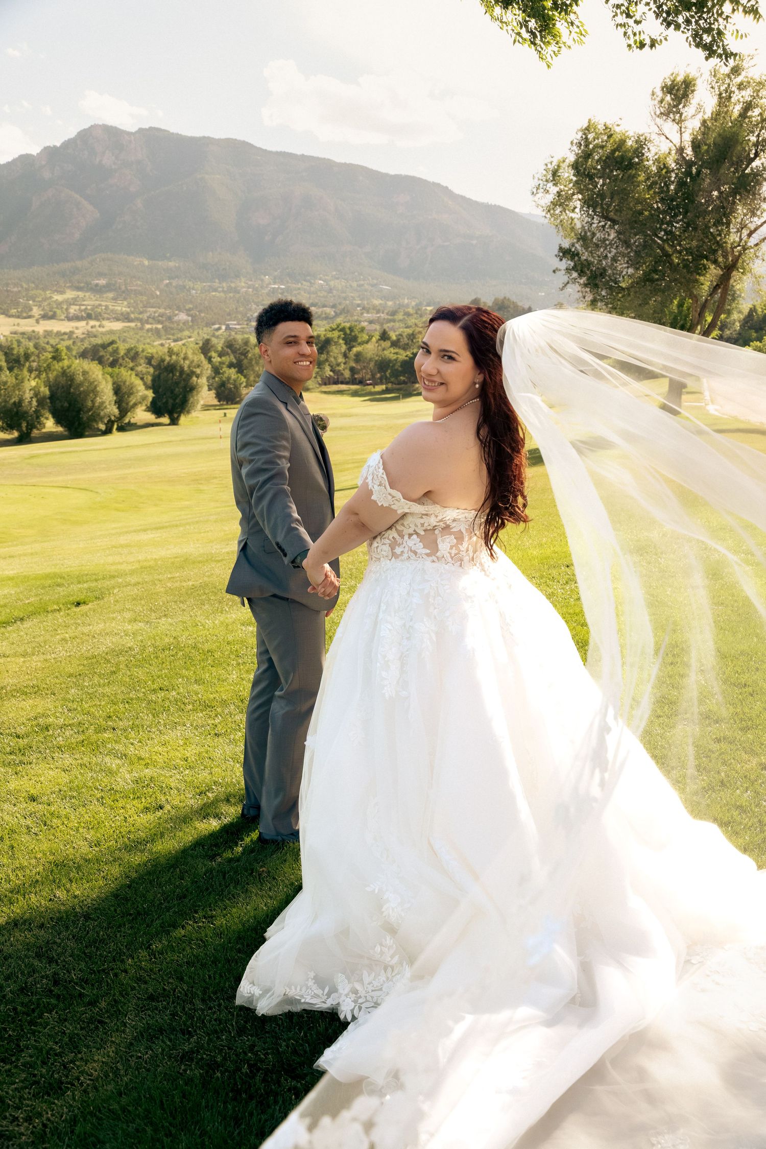 Bride in white lace gown with flowing veil holds groom's hand on green lawn with mountain backdrop.