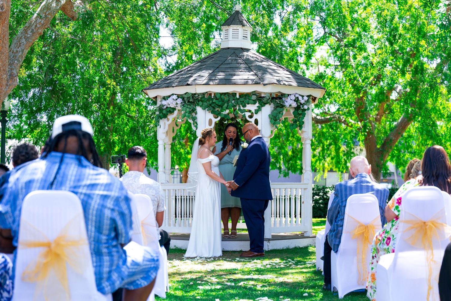 A garden themed wedding ceremony in progress. The bride in a white dress and groom in a blue suit hold hands at the altar. Wedding photo LA
