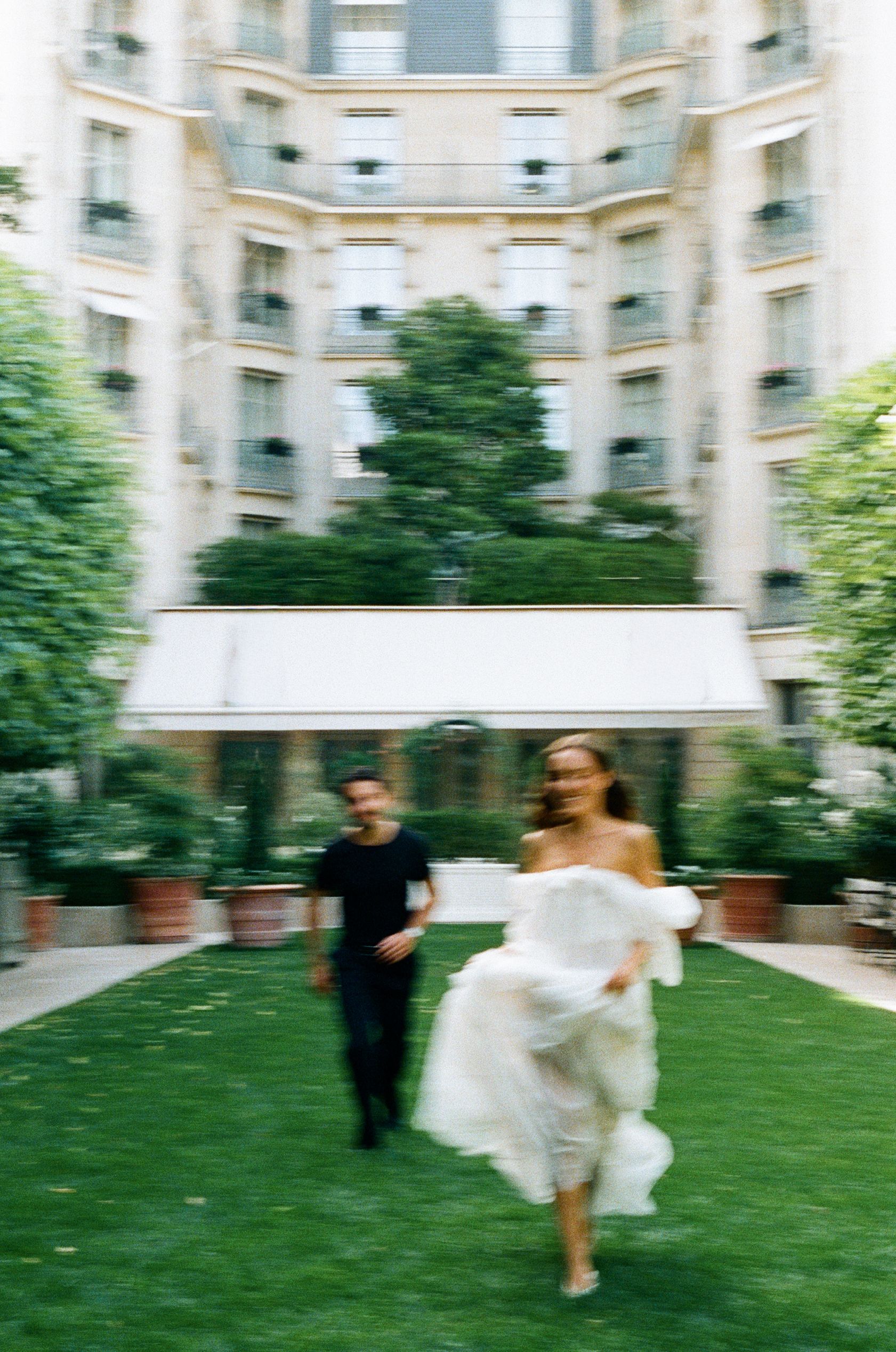 Bride in white dress runs across lush garden courtyard with companion, elegant Parisian building backdrop.