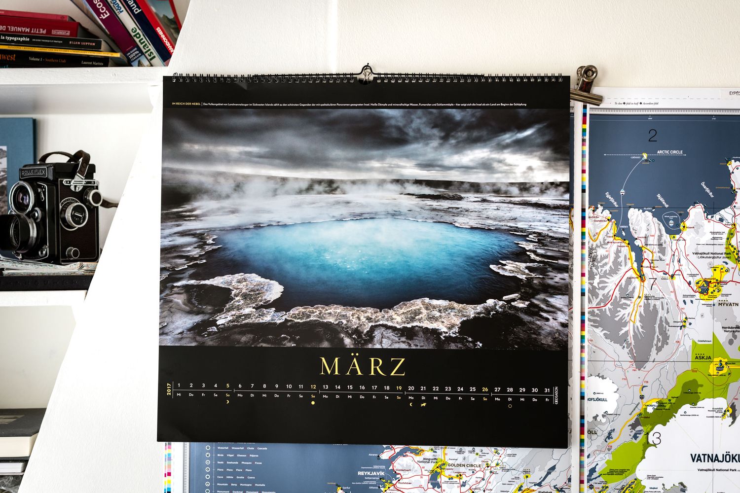 A March wall calendar featuring a stunning blue geothermal hot spring in Iceland, displayed above a bookshelf with a vintage camera.
