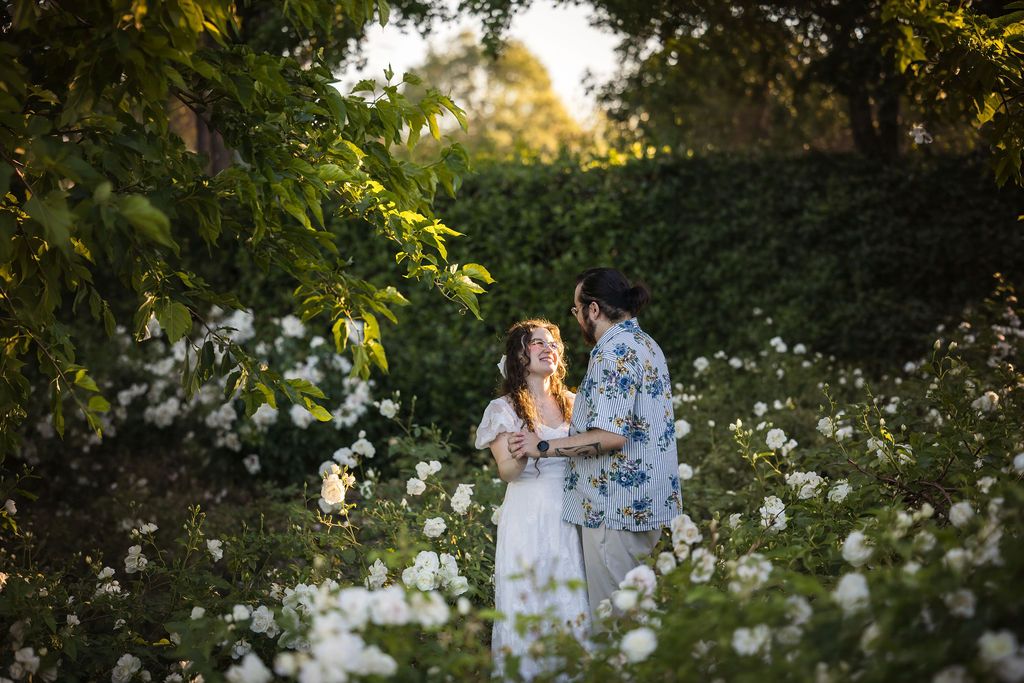 Couple sharing a romantic moment surrounded by white flowers and lush greenery in golden hour light.