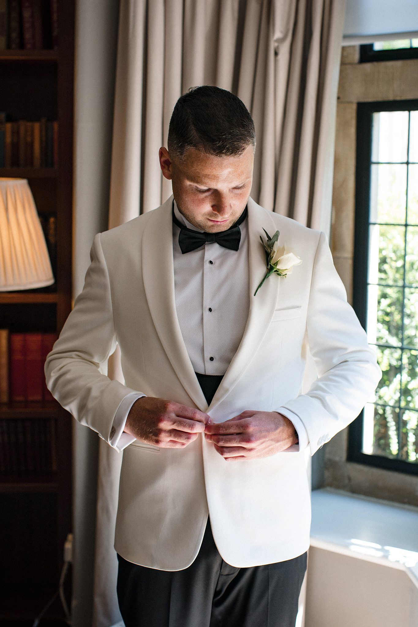 Groom in white tuxedo jacket with black bow tie and boutonniere buttoning his jacket before wedding ceremony.