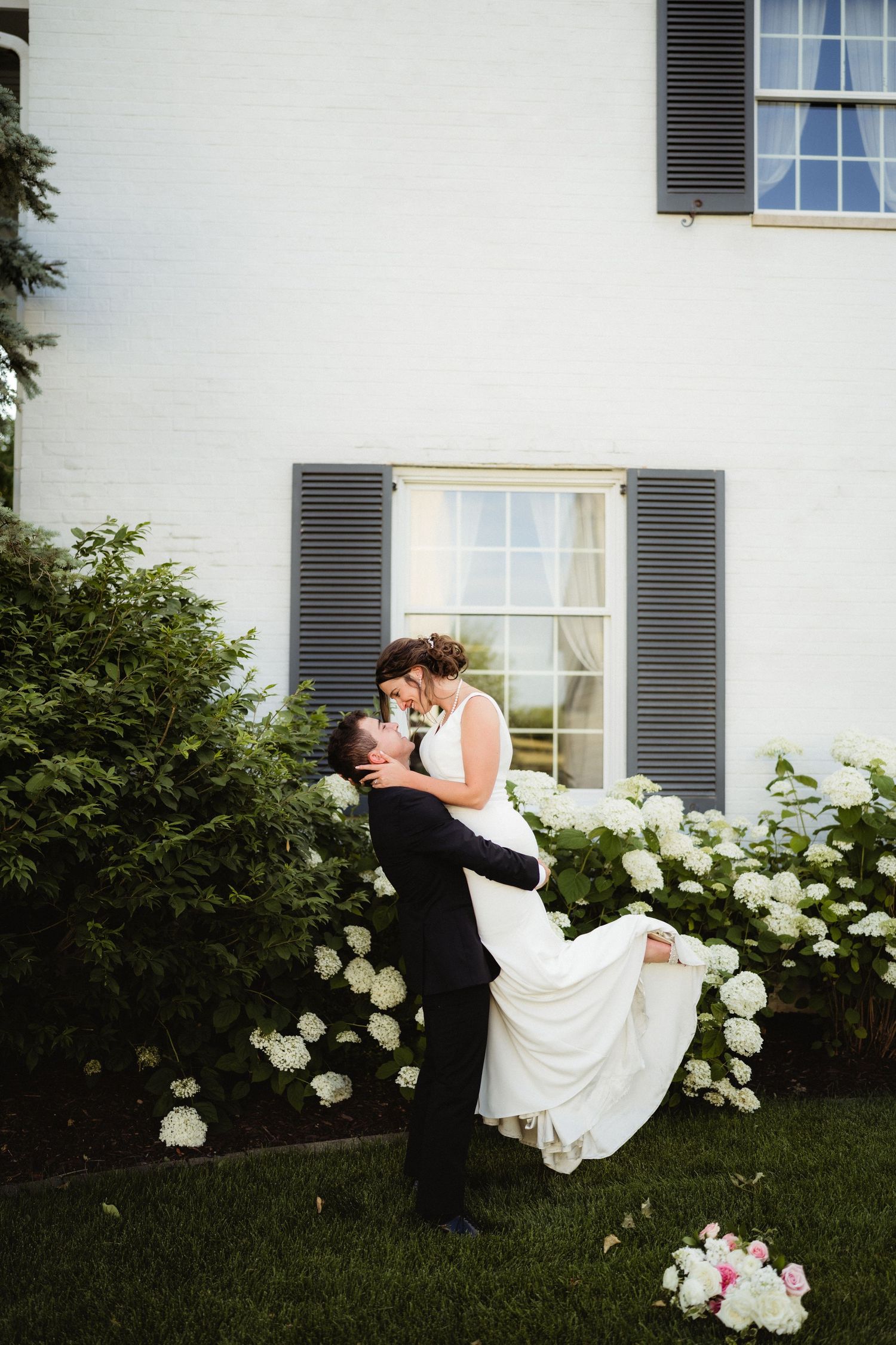 Groom lifts bride in white dress outside white house with gray shutters and blooming hydrangeas on wedding day.