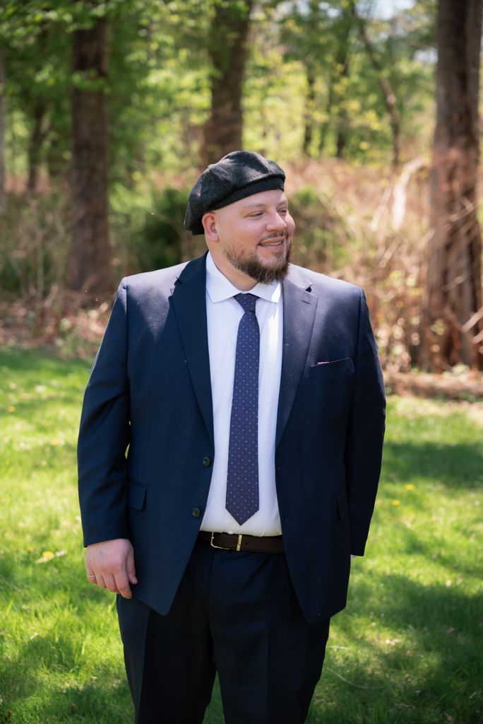 Man in navy suit, blue tie, and black beret smiling outdoors in a green wooded setting.