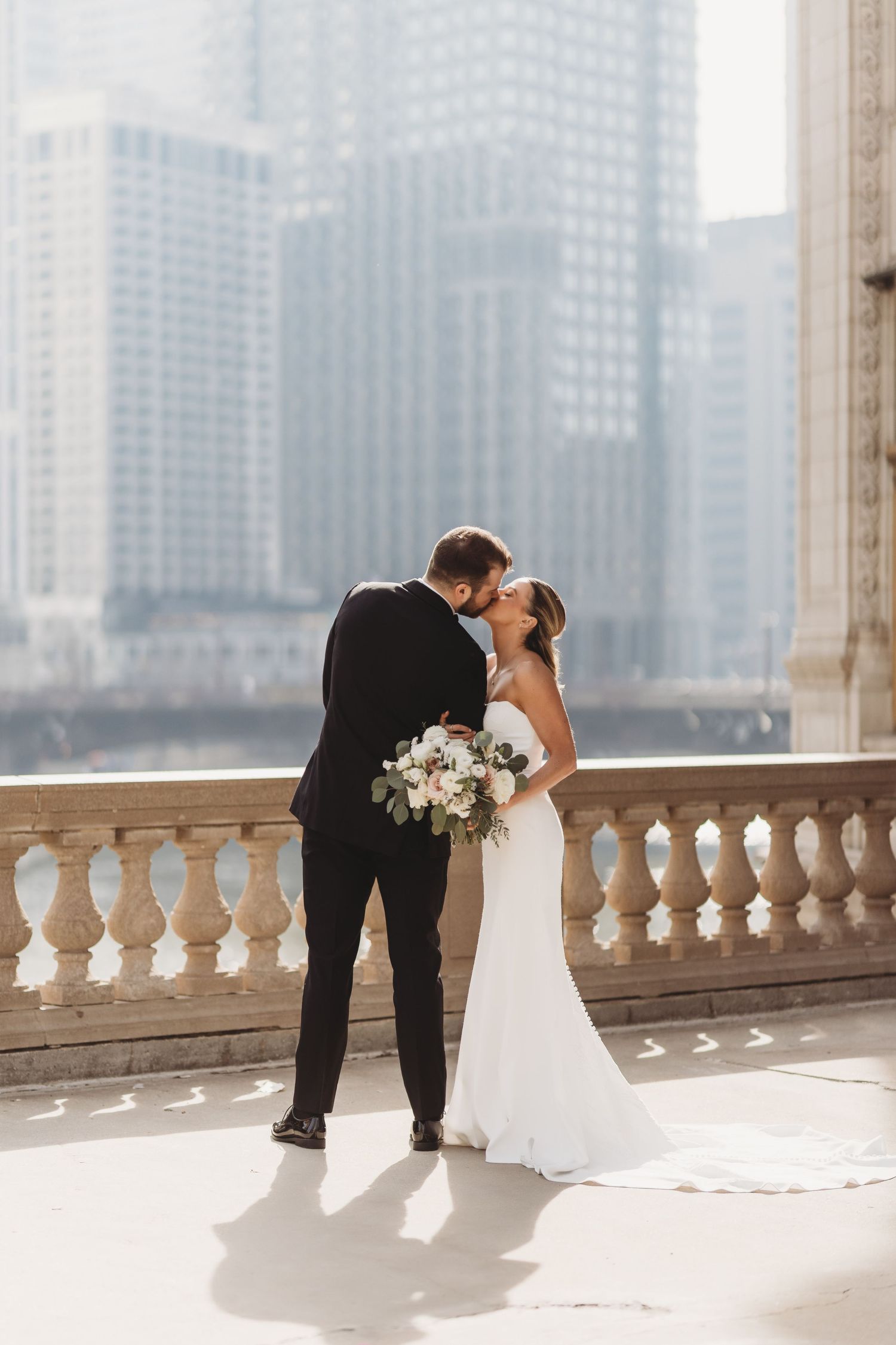 A bride and groom share a romantic moment on a city balcony, with the bride holding a white floral bouquet.