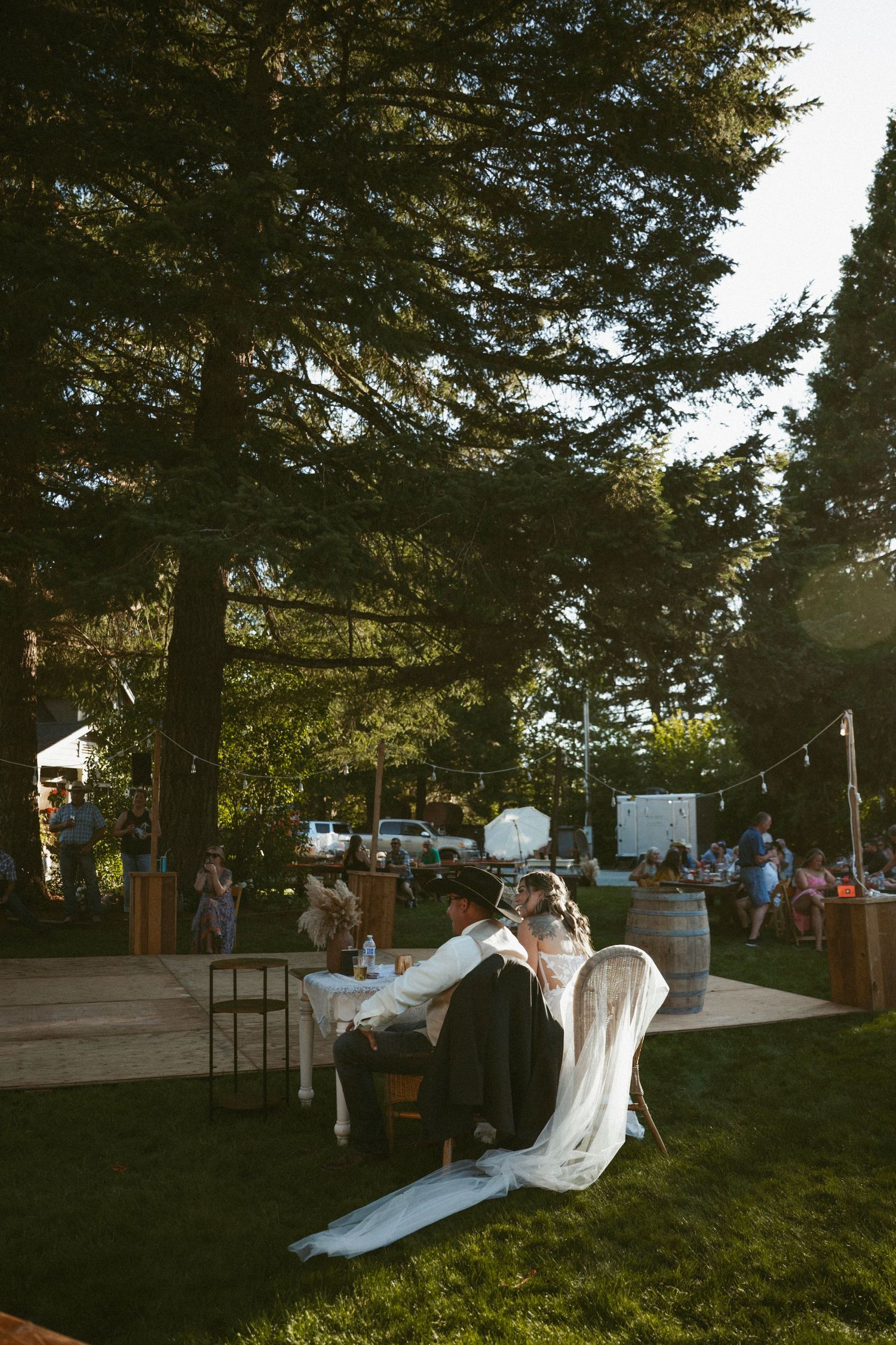 Bride and groom sit together at sweetheart table during outdoor wedding reception surrounded by tall pine trees.