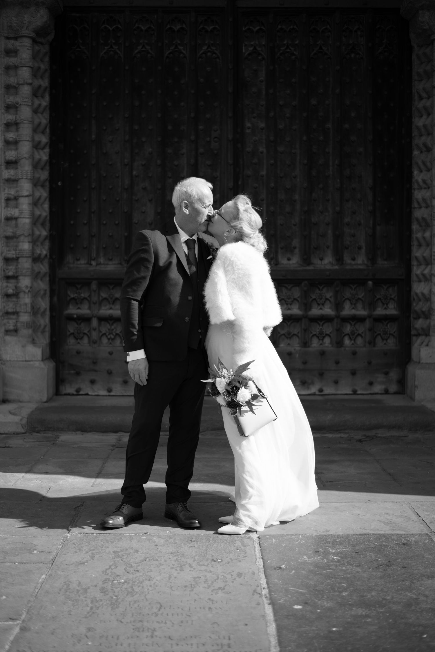 Black and white photo of a bride and groom sharing a kiss outside an ornate church door on their wedding day.