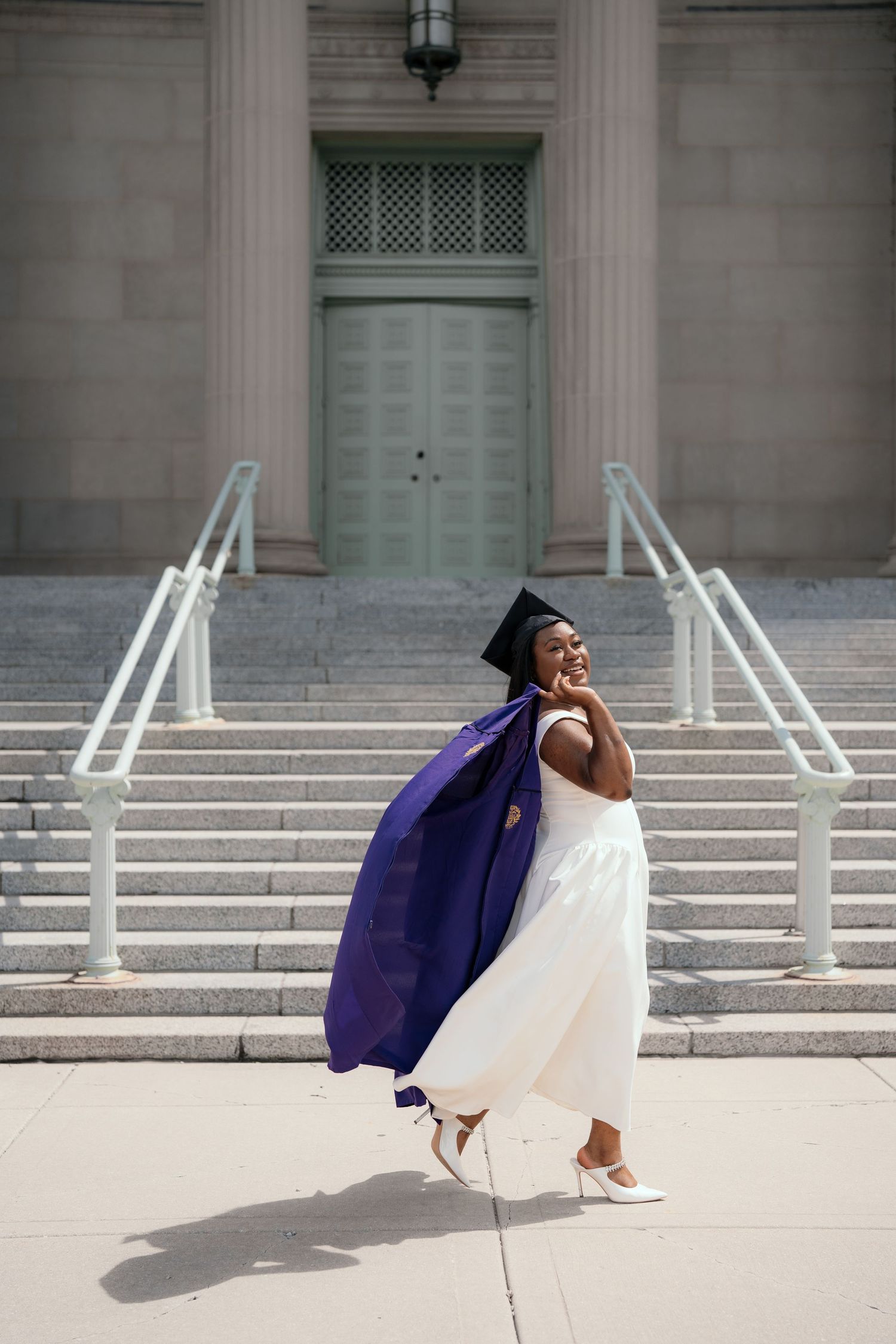 Joyful graduate in white dress and purple cap and gown celebrating on university steps.