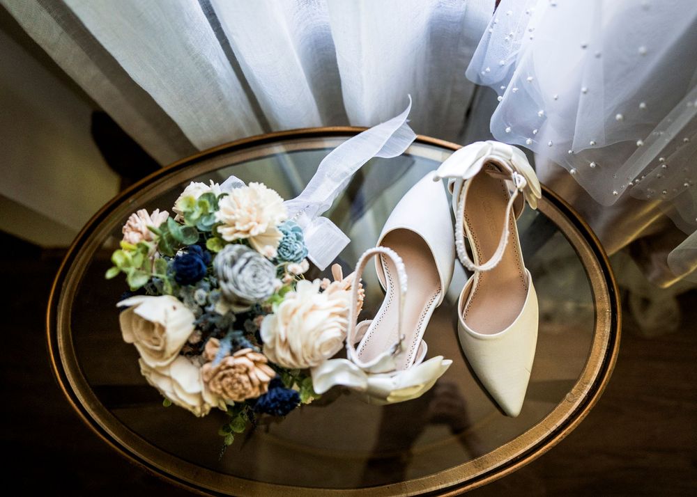 Bridal heels and floral bouquet with blue and white flowers on a round glass table near a wedding dress.