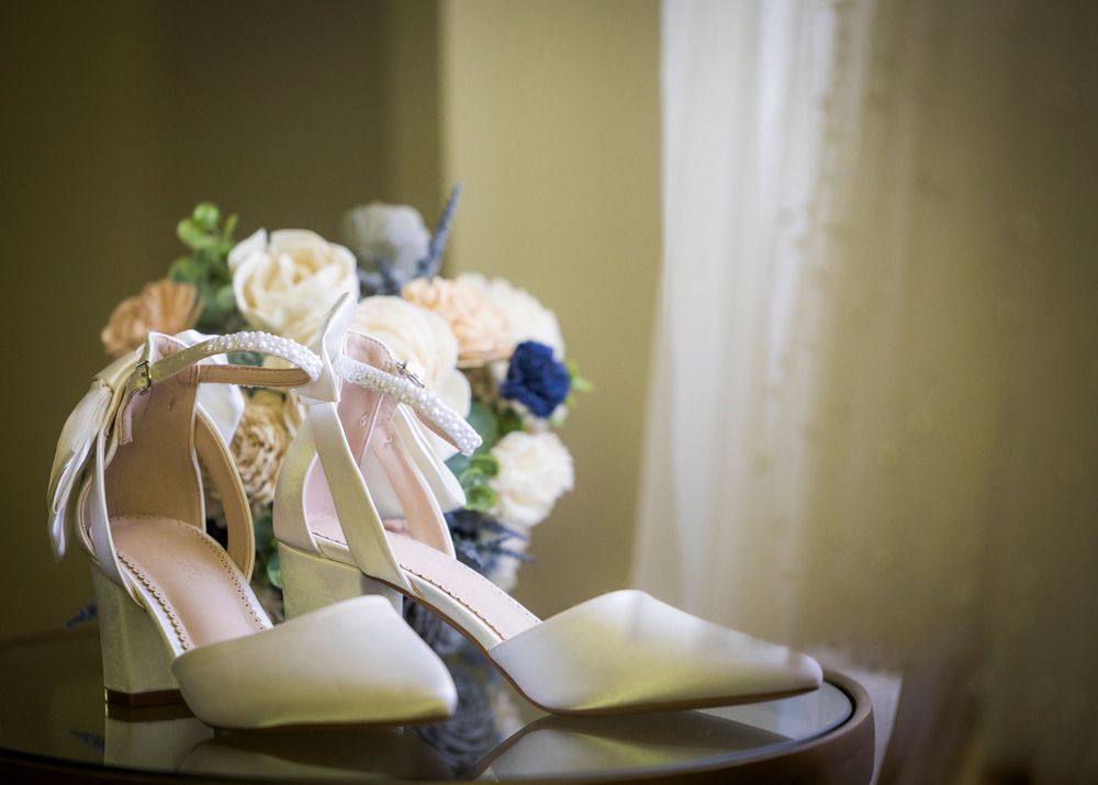 Elegant white bridal heels with ankle straps displayed on a table with a floral bouquet in soft blue and pink tones.