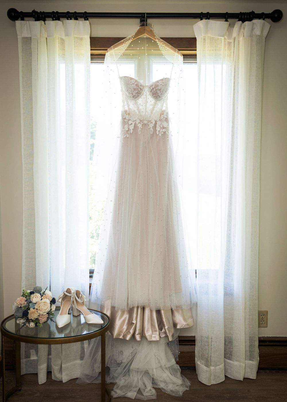 Elegant strapless lace wedding gown hanging by window with bouquet and heels.
