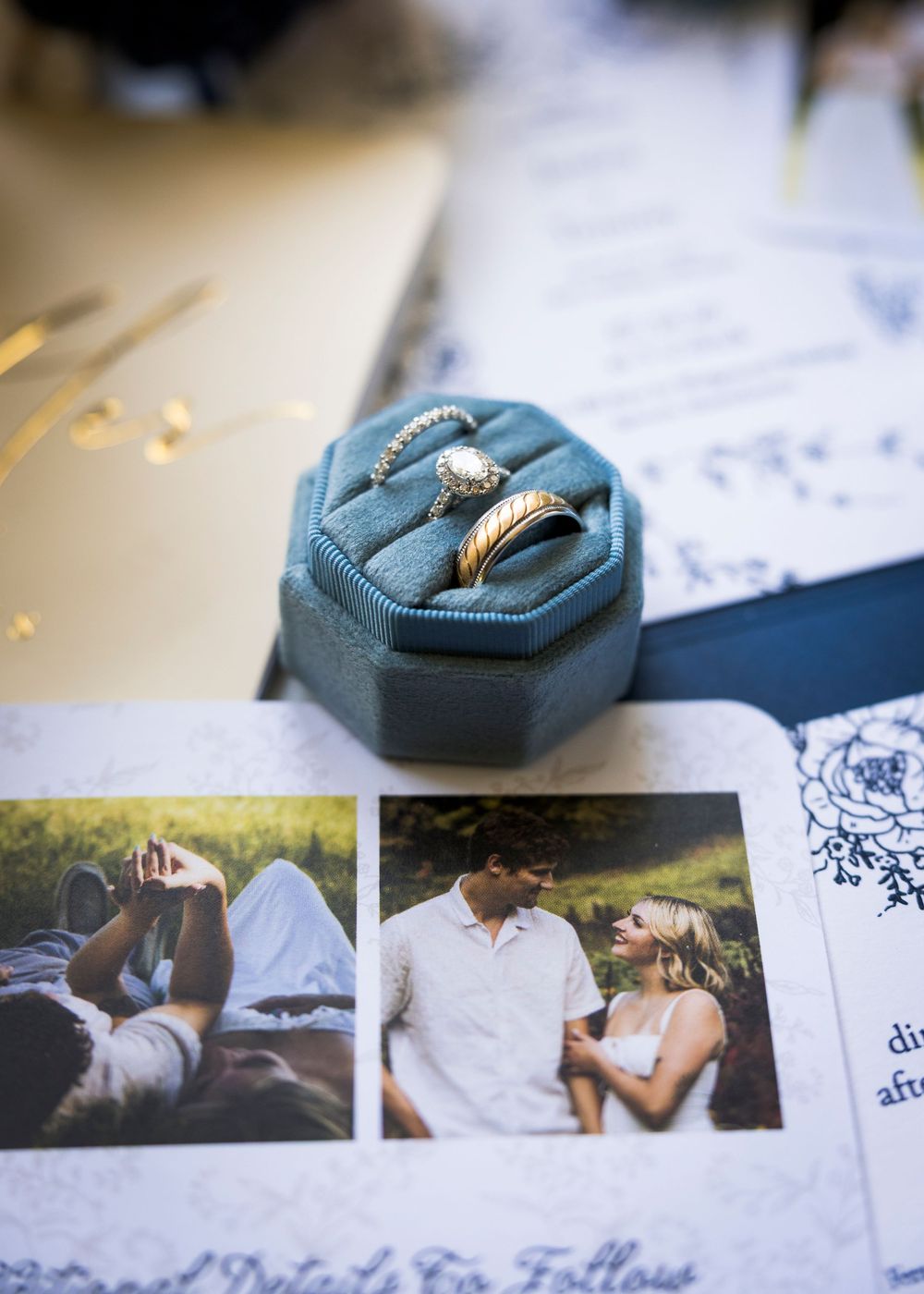Close-up of wedding rings in a teal velvet octagonal ring box atop navy blue wedding invitation suite with couple photos.