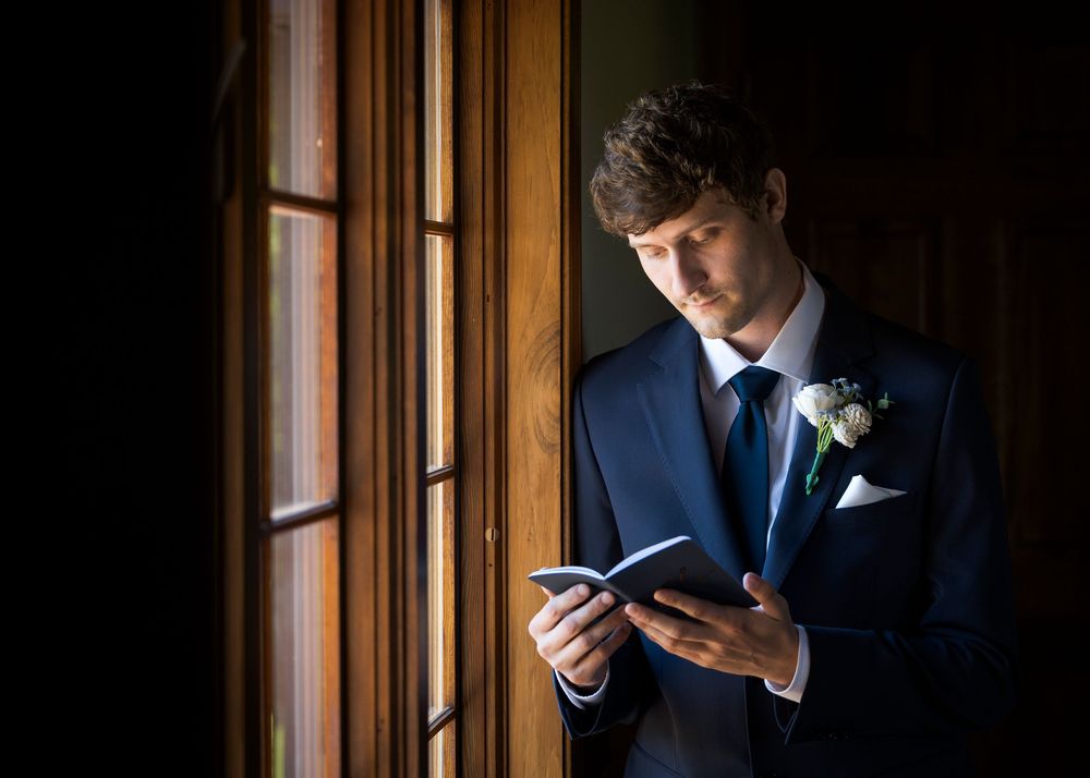 Handsome groom in navy suit reading vows by wooden window before ceremony.