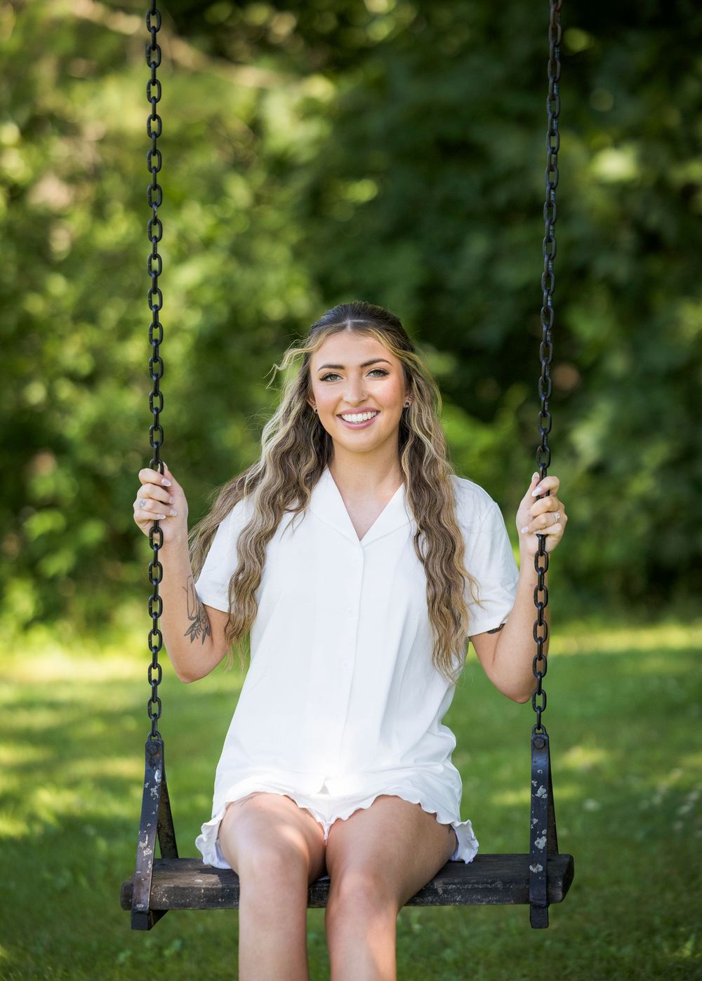 Young woman in white dress smiling on a wooden swing surrounded by lush green trees outdoors.