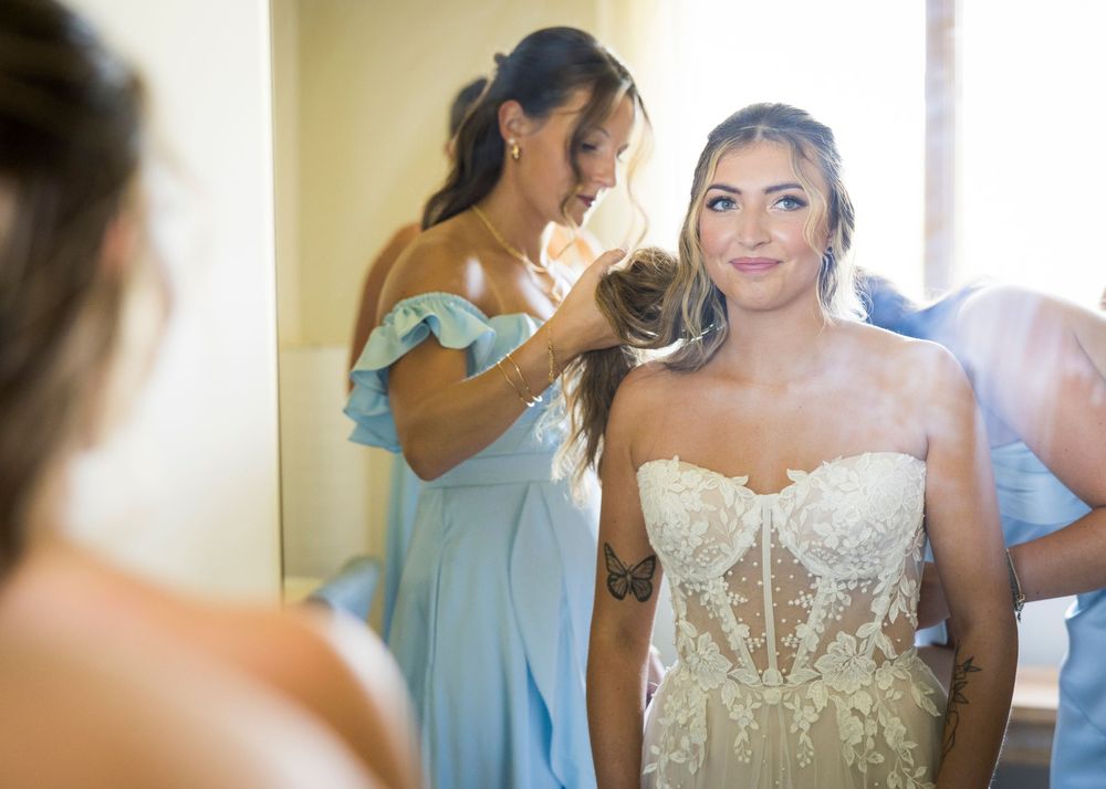 Bride in lace strapless wedding gown getting hair styled by bridesmaid in blue dress before ceremony.