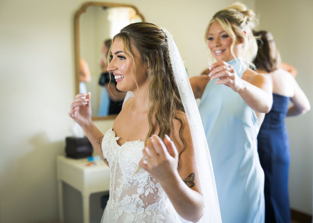 Bride smiling in lace gown while bridesmaids help with veil preparations.