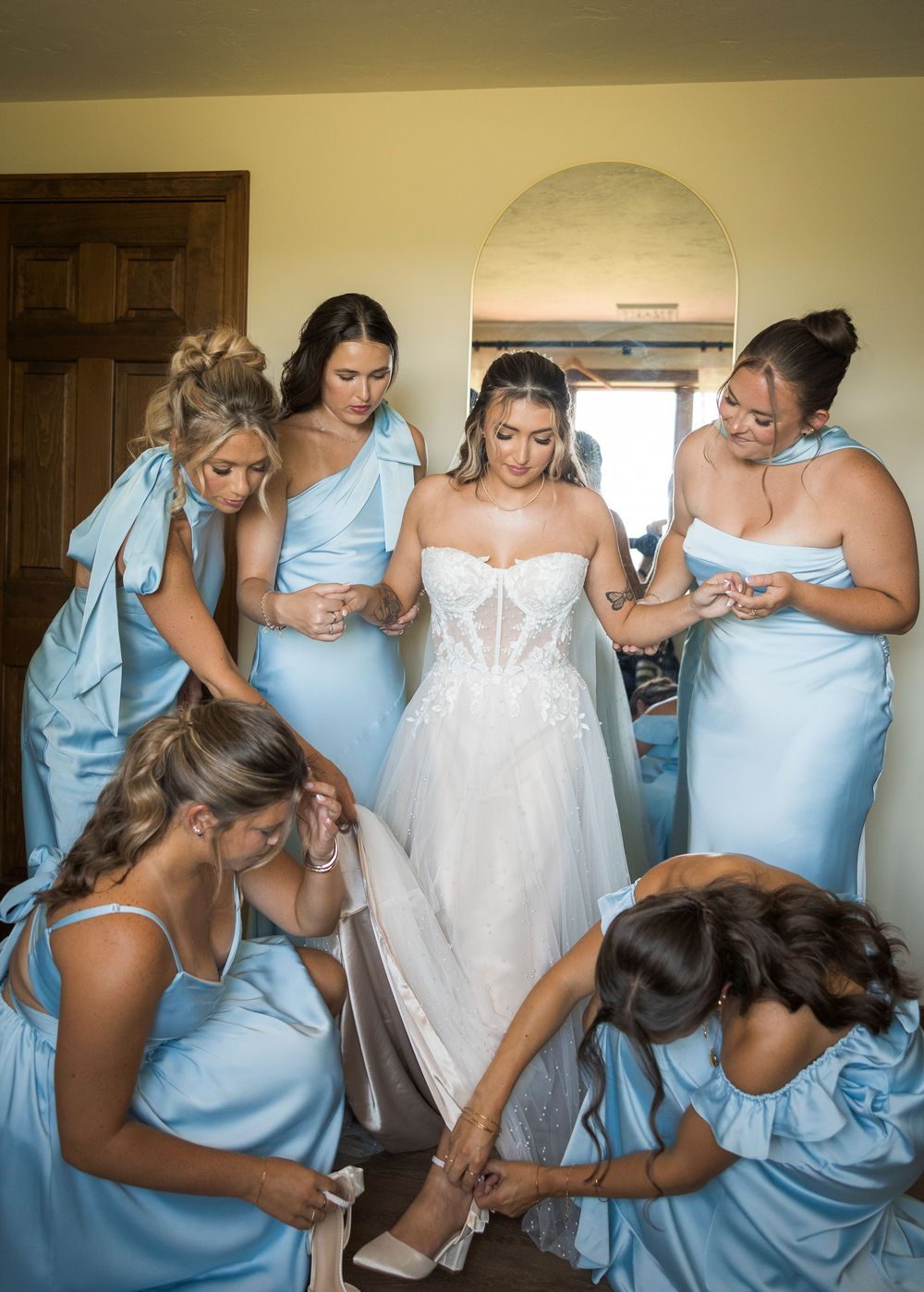 Bride in white lace gown surrounded by bridesmaids in blue satin dresses helping her get ready.