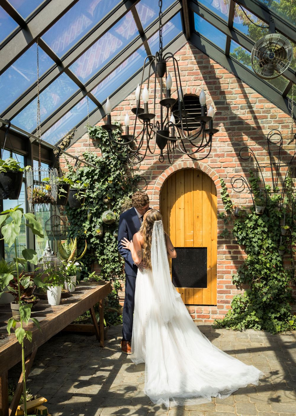 Bride and groom share a romantic moment in a charming greenhouse with brick walls, lush greenery, and a yellow arched door.