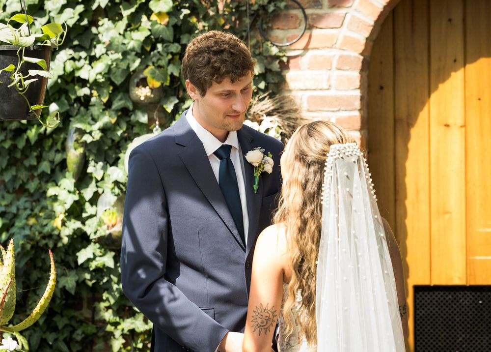 Groom in navy suit gazing at bride in veil during outdoor wedding ceremony near brick wall.