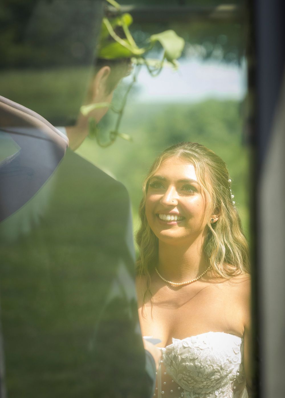 Bride smiling at groom during outdoor wedding ceremony, wearing white strapless gown and pearl necklace.