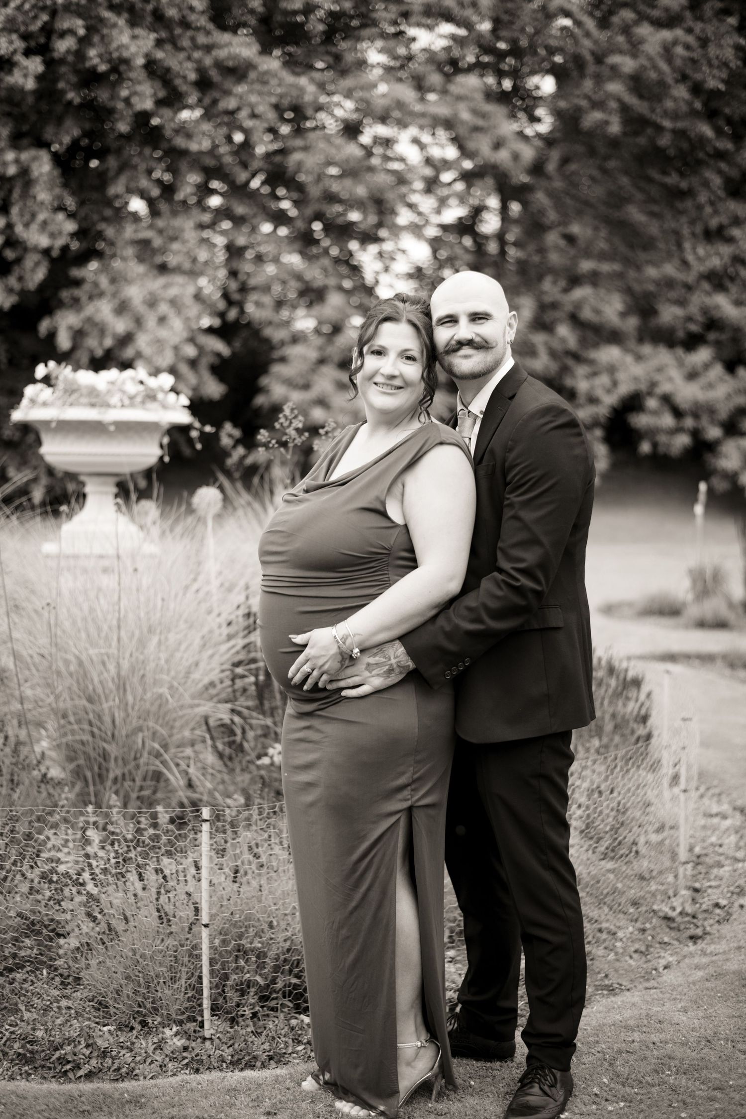 Black and white photo of a couple posing in a garden, man in suit embracing woman in elegant gown.