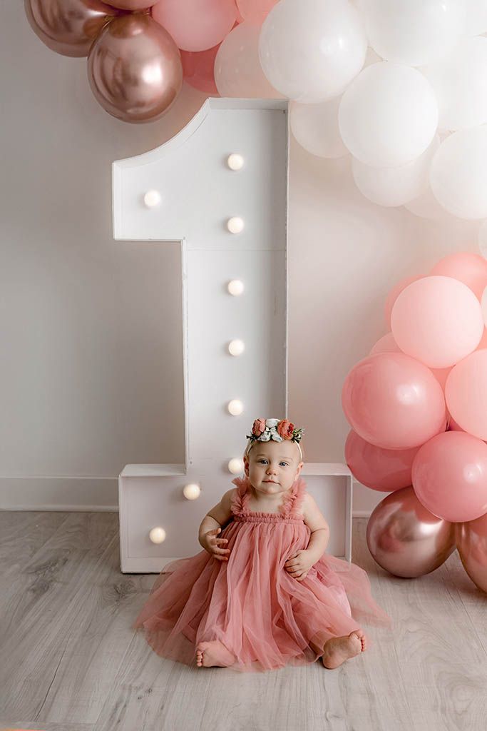Baby girl in pink tutu dress celebrating first birthday with balloon arch and marquee number one.