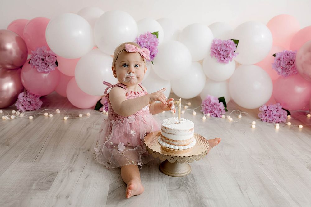 Baby girl in pink tutu smashes first birthday cake surrounded by pink and white balloons and flowers.