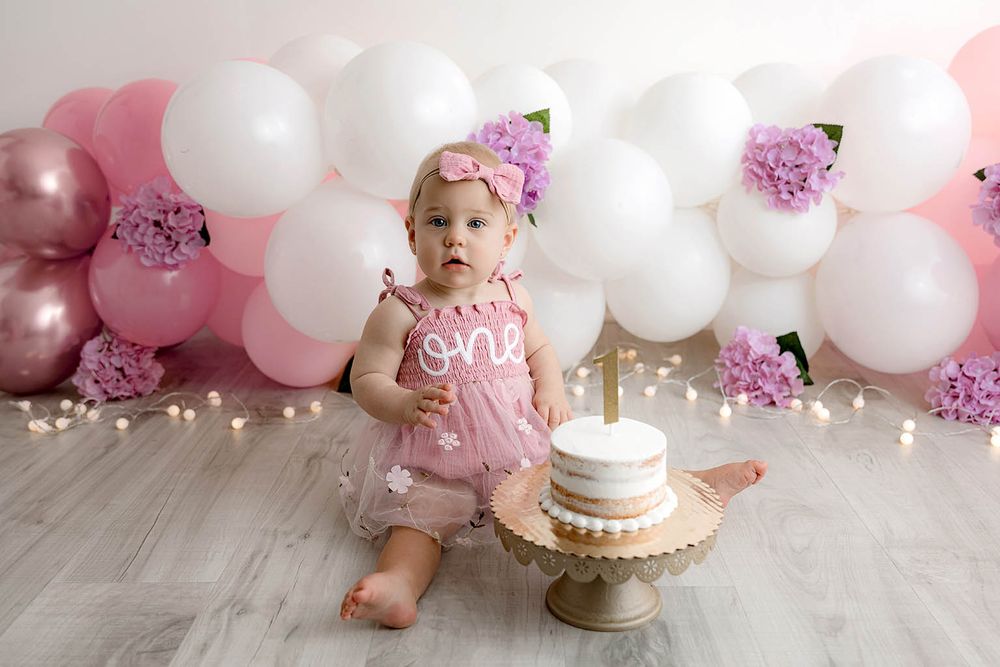 Baby girl in pink dress celebrating first birthday with cake smash, surrounded by pink and white balloons.