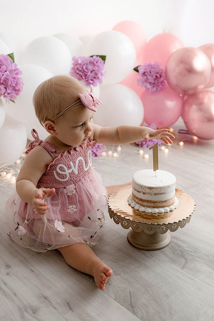 Baby girl in pink tutu dress reaching for her first birthday cake surrounded by balloons.