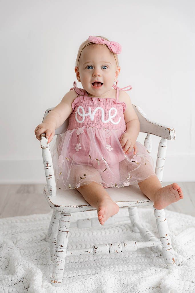Smiling baby girl in pink 'one' romper sitting in a rustic white chair for her first birthday photo.