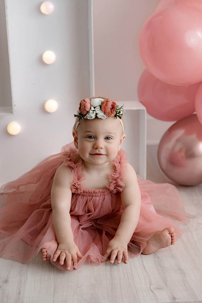 Adorable baby girl in pink tulle dress wearing floral crown smiling at her first birthday photoshoot.