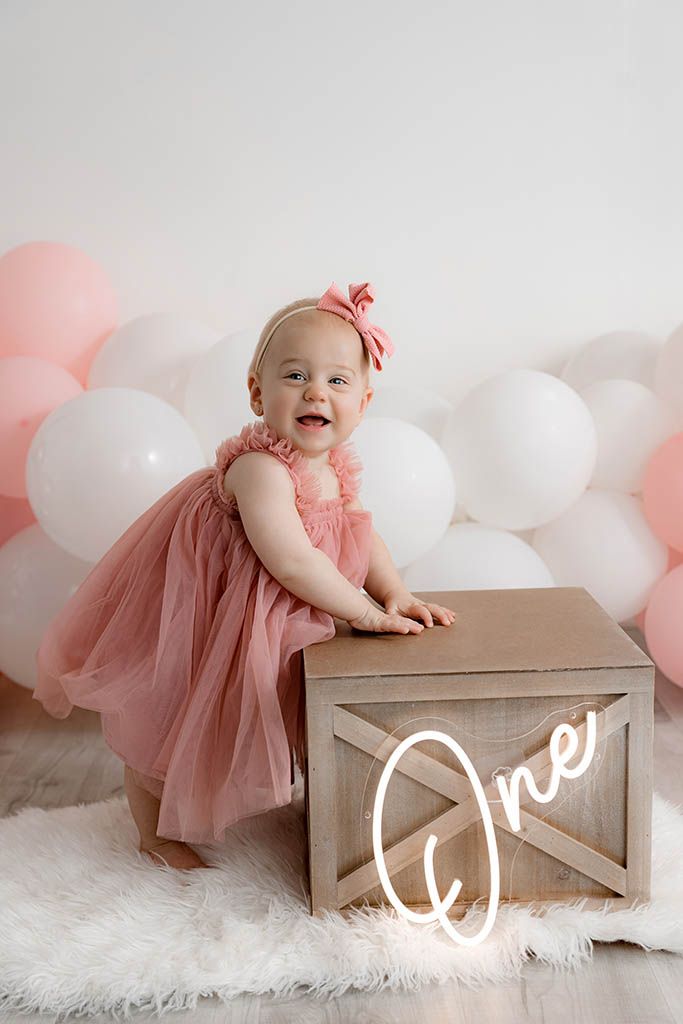 Happy baby girl in pink dress standing at a wooden crate with One neon sign and balloons.