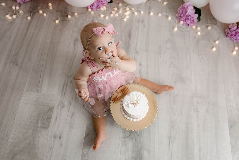 Baby girl in pink romper sits on floor with smash cake, balloons, and floral decor for first birthday photo.