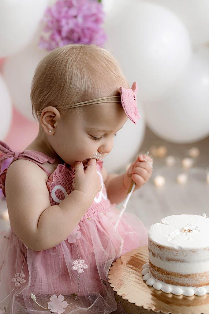 Baby girl in pink dress tasting her first birthday smash cake surrounded by balloons and flowers.