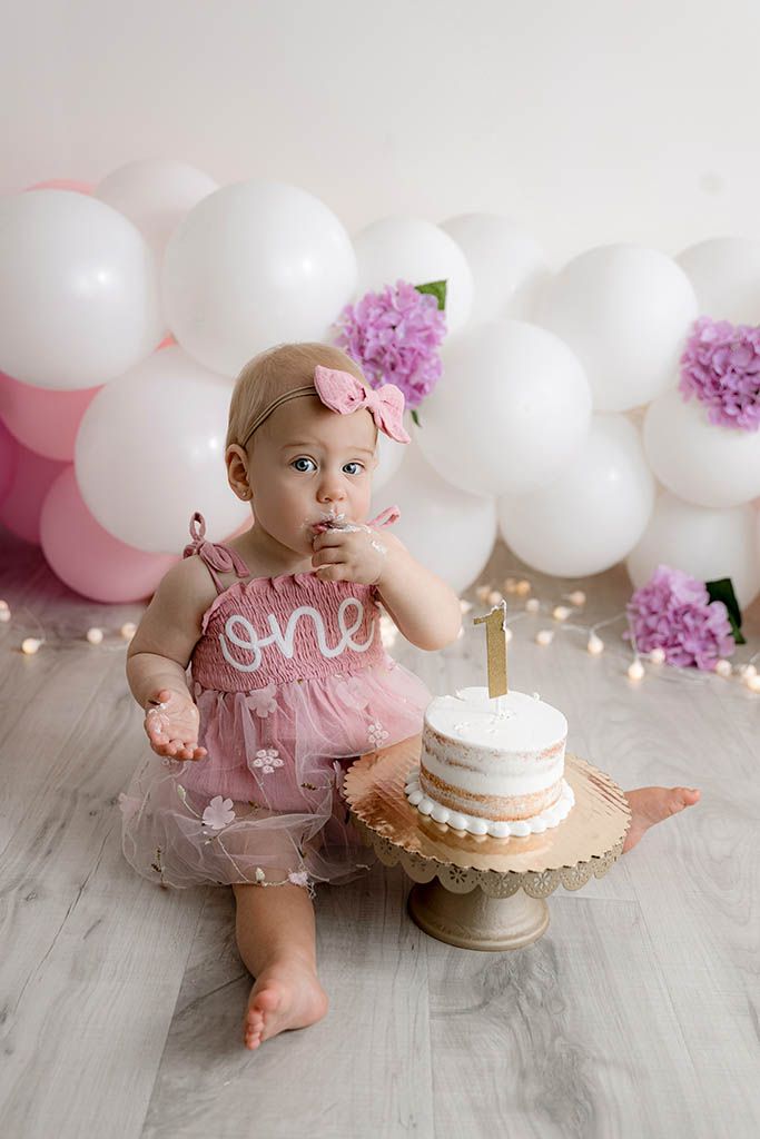 Baby girl in pink One romper eating cake at her first birthday smash cake photoshoot with balloons.