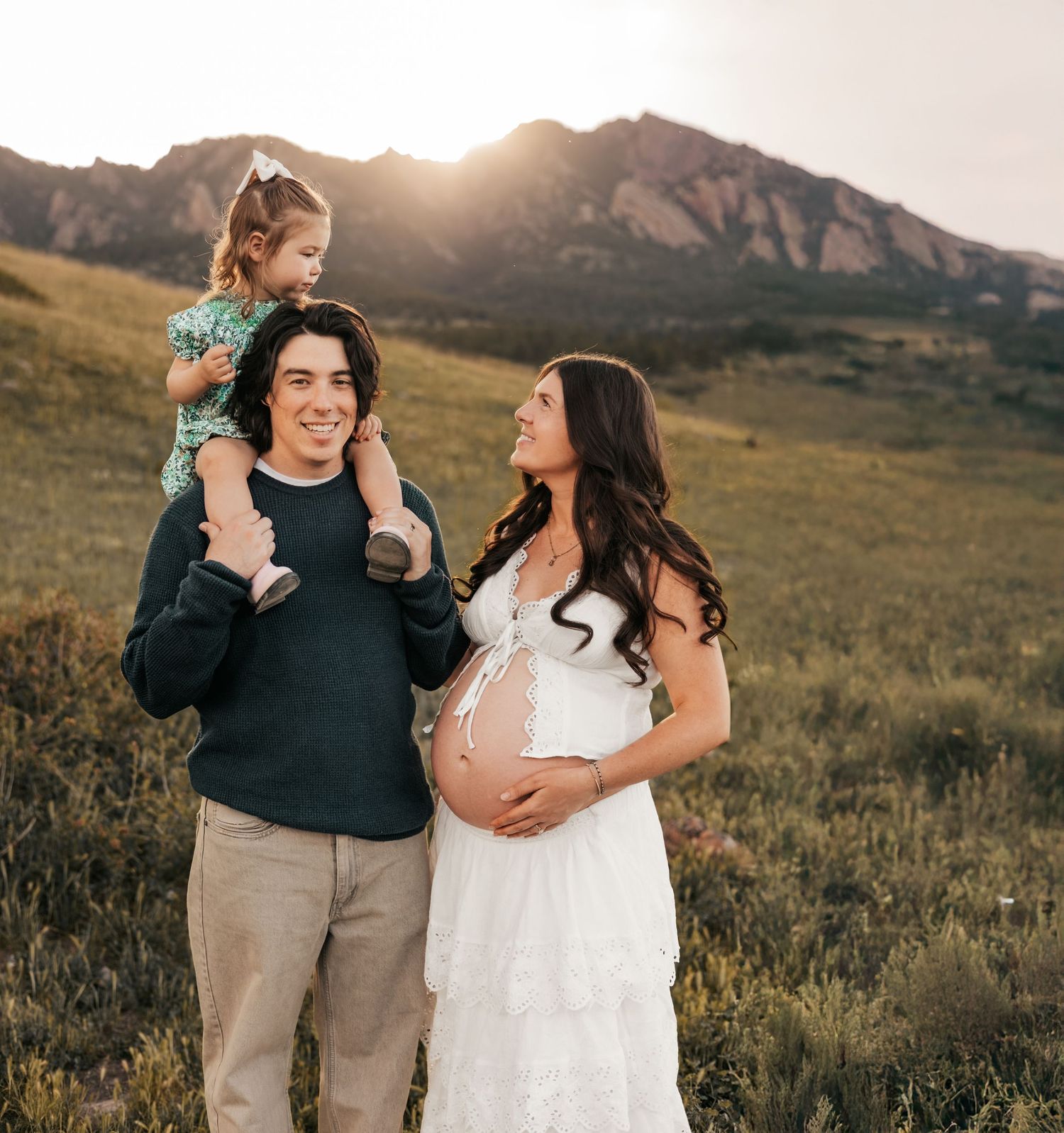 Pregnant woman in white dress with partner and toddler on shoulders posing in mountain meadow at golden hour | Boulder family photograher